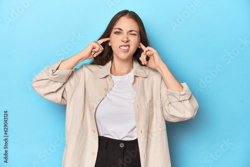 Stylish young woman in an overshirt on a blue background covering ears with fingers, stressed and desperate by a loudly ambient.