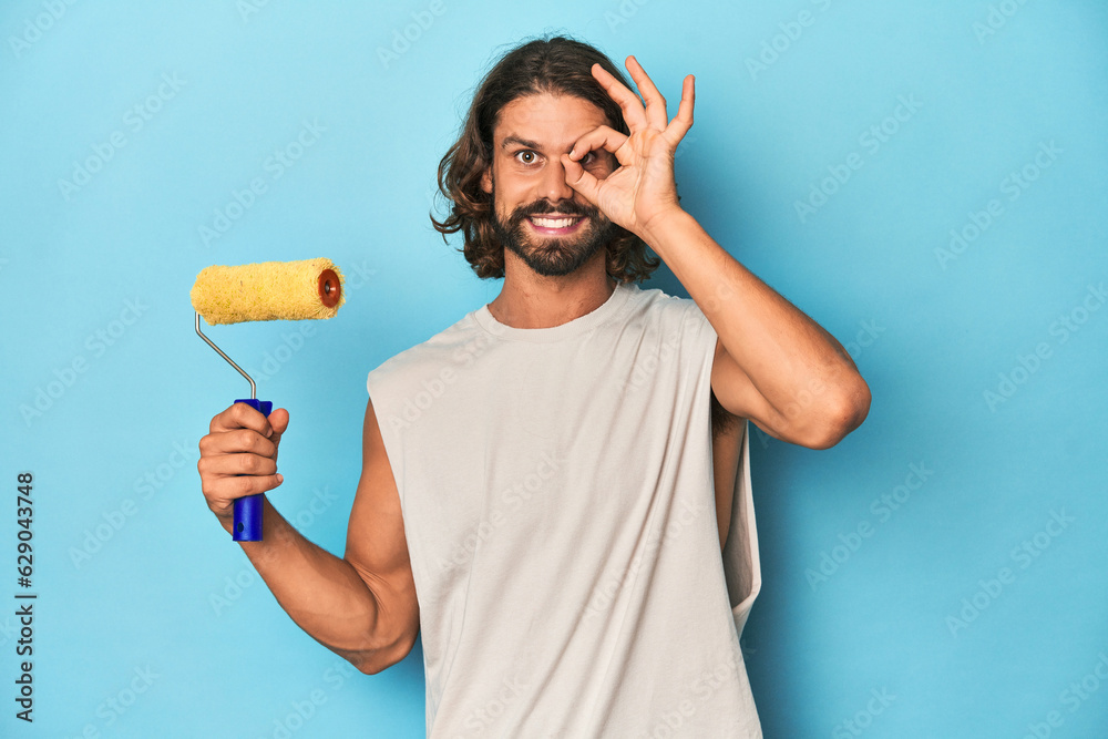 Bearded man painting with a yellow roller excited keeping ok gesture on eye. Bearded man painting with a yellow roller excited keeping ok gesture on eye.