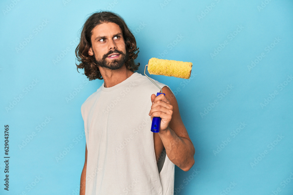 Bearded man painting with a yellow roller looks aside smiling, cheerful and pleasant.