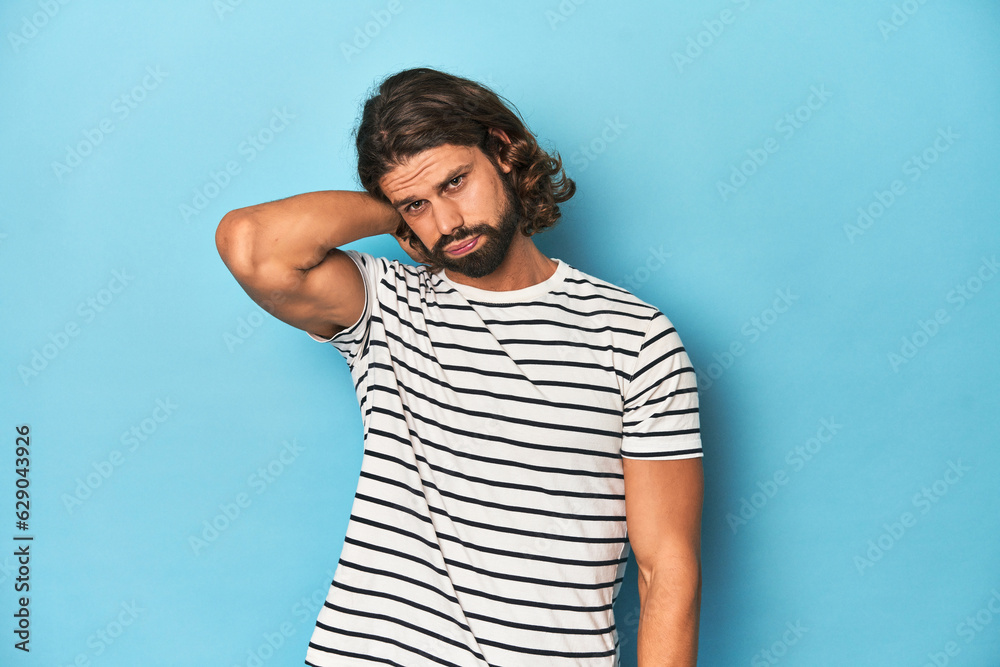 Bearded man in a striped shirt, blue backdrop suffering neck pain due to sedentary lifestyle. Bearded man in a striped shirt, blue backdrop suffering neck pain due to sedentary lifestyle.