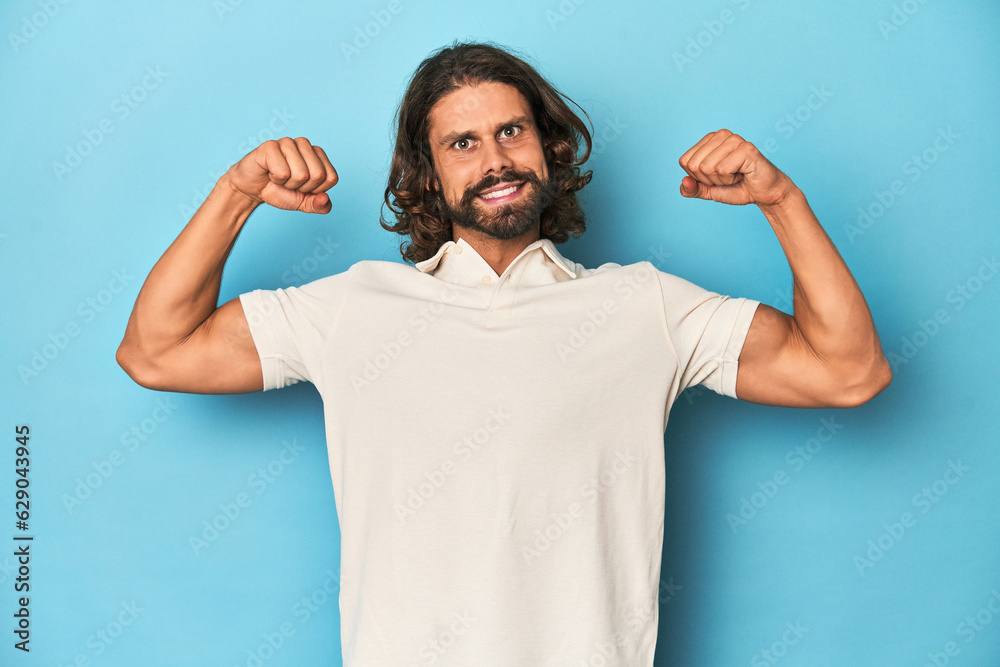 Long-haired man in a white polo, blue studio showing strength gesture with arms, symbol of feminine power Long-haired man in a white polo, blue studio showing strength gesture with arms, symbol of feminine power