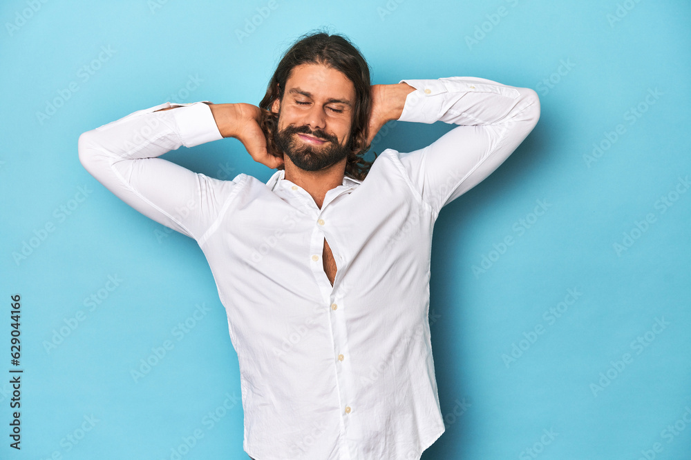 Bearded man in a white shirt, blue backdrop feeling confident, with hands behind the head.