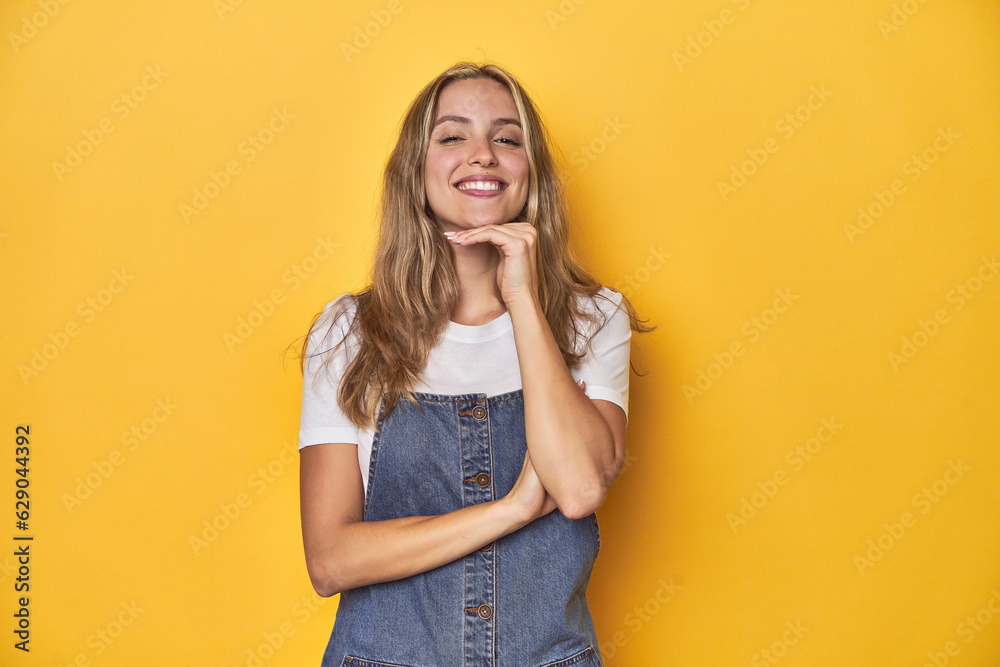 Young blonde Caucasian woman in denim overalls posing on a yellow background, smiling happy and confident, touching chin with hand.