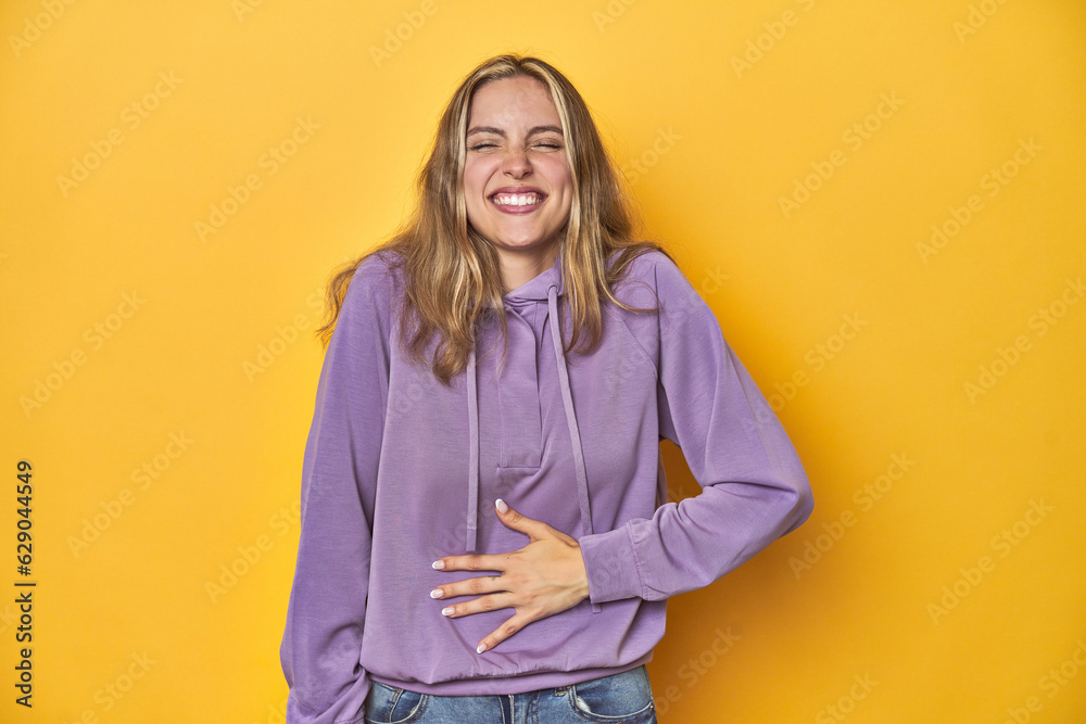 Young blonde Caucasian woman in a violet sweatshirt on a yellow background, touches tummy, smiles gently, eating and satisfaction concept. Young blonde Caucasian woman in a violet sweatshirt on a yellow background, touches tummy, smiles gently, eating and satisfaction concept.