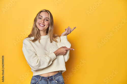 Fototapete Young blonde Caucasian woman in a white sweatshirt on a yellow studio background, excited pointing with forefingers away