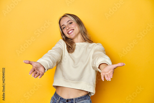 Young blonde Caucasian woman in a white sweatshirt on a yellow studio background  showing a welcome expression.