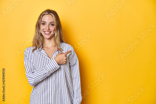 Young blonde Caucasian woman in a striped business shirt on a yellow background, smiling and pointing aside, showing something at blank space. © Asier