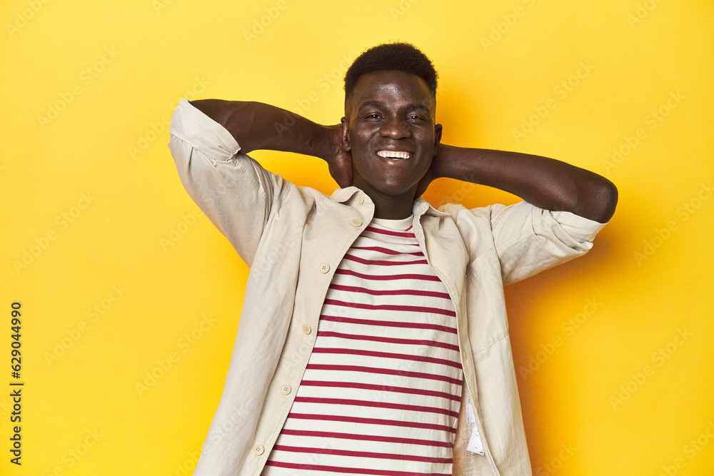 Stylish young African man on vibrant yellow studio background, feeling confident, with hands behind the head.