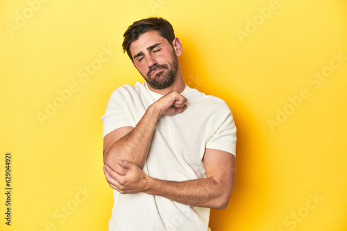 Caucasian man in white t-shirt on yellow studio background massaging elbow, suffering after a bad movement.