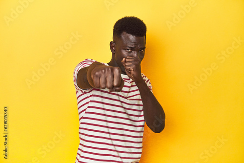 Stylish young African man on vibrant yellow studio background, throwing a punch, anger, fighting due to an argument, boxing.