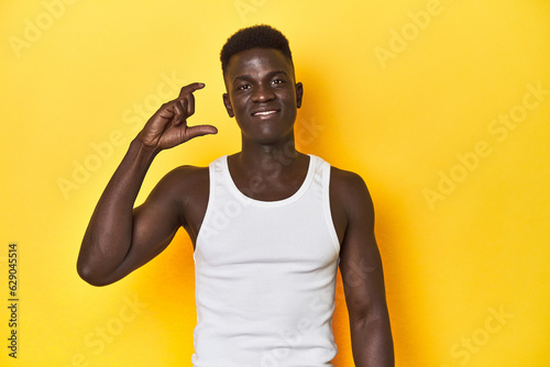 Stylish young African man on vibrant yellow studio background, holding something little with forefingers, smiling and confident.