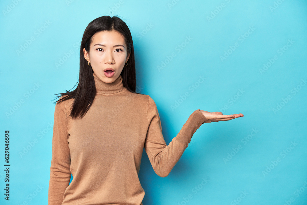 Young Asian woman in brown turtleneck, impressed holding copy space on palm. Young Asian woman in brown turtleneck, impressed holding copy space on palm.