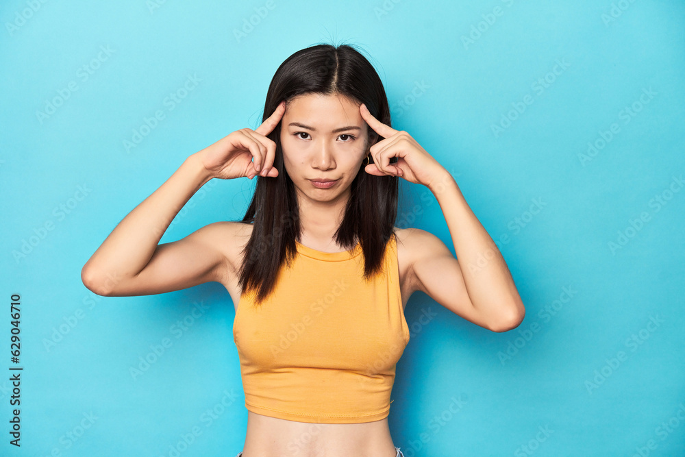 Asian woman in summer yellow top, studio setup, focused on a task, keeping forefingers pointing head.