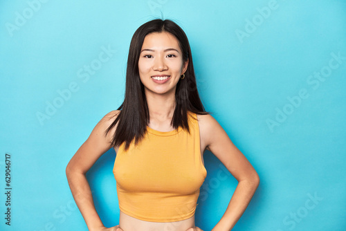 Asian woman in summer yellow top, studio setup, confident keeping hands on hips.