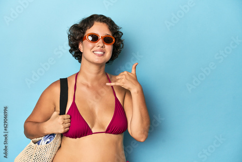 Young woman in bikini with beach bag showing a mobile phone call gesture with fingers. © Asier