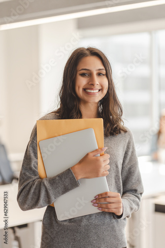 Smiling confident Indian business woman holding documents looking at camera while standing at modern office. Successful business concept