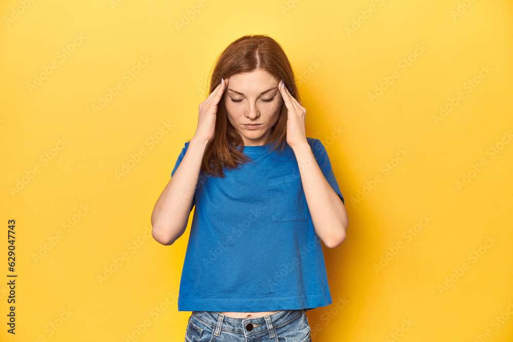 Redhead in blue t-shirt on yellow backdrop touching temples and having headache.