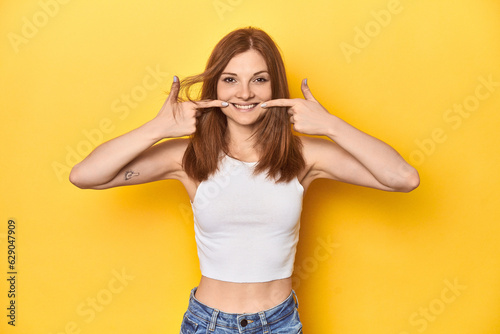 Redhead in white tank top, relaxed studio pose smiles, pointing fingers at mouth.