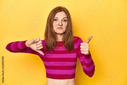 Redhead in pink-red striped shirt, casual look showing thumbs up and thumbs down, difficult choose concept