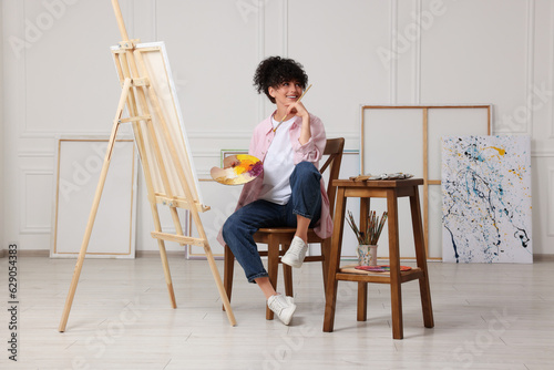 Young woman holding brush and artist`s palette near easel with canvas