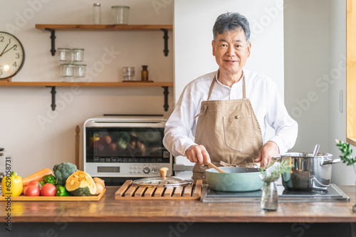 家のキッチンでフライパンを使って手料理を作るアジア人の高齢者男性（笑顔）
