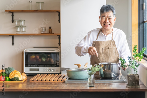 家のキッチンで鍋を使って煮込み料理を作るアジア人の高齢者男性（笑顔）

