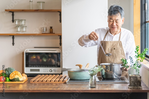 Fotografiet 家のキッチンで料理を作って味見するアジア人の高齢者男性（笑顔）
