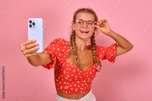 Young laughing Caucasian woman adjusting fashion glasses and making video call on mobile phone chatting with friends from college or university via internet stands on pink background.