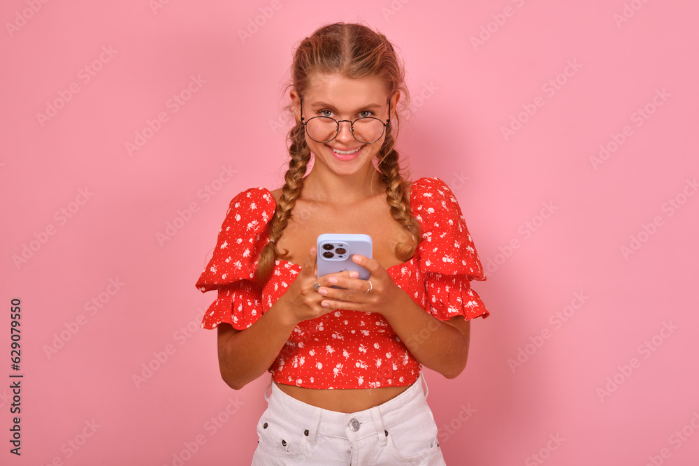 Young cheerful beautiful Caucasian woman student in glasses holding mobile phone getting distance education and studying in internet institute or college through website stands in pink studio. Young cheerful beautiful Caucasian woman student in glasses holding mobile phone getting distance education and studying in internet institute or college through website stands in pink studio.