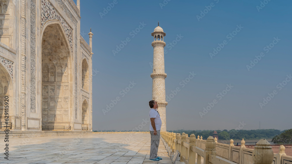 A man stands next to the Taj Mahal at the fence, looking into the distance. The hand is raised to the eyes. A white marble mausoleum and a high minaret against the blue sky. India. Agra.