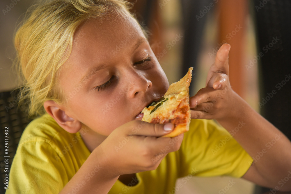 Adorable curly blonde boy eating pizza at the camera in italian restaurant. Portrait of child with long blonde hair and a yellow T-shirt eats junk food in a cafe. Blur on the background. Adorable curly blonde boy eating pizza at the camera in italian restaurant. Portrait of child with long blonde hair and a yellow T-shirt eats junk food in a cafe. Blur on the background.
