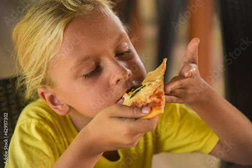 Adorable curly blonde boy eating pizza at the camera in italian restaurant. Portrait of child with long blonde hair and a yellow T-shirt eats junk food in a cafe. Blur on the background.