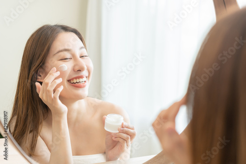 Facial beauty skin care, smile of pretty asian young woman in bathrobe looking at mirror, hand applying moisturizer lotion on her face, holding jar of skin cream before makeup cosmetic routine at home