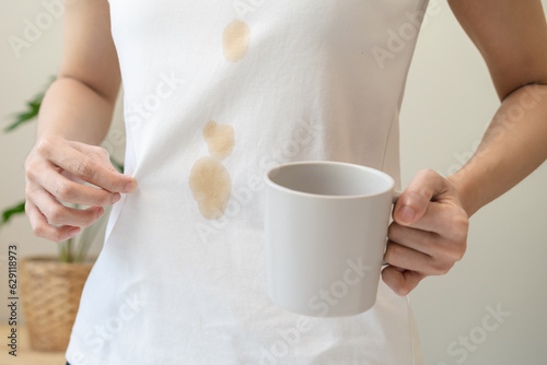 Cloth stain, disappointment asian young woman clumsy with hot coffee, tea stains on shirt, hand show making spill drop on white t-shirt, spot dirty or smudge on clothes at home, isolated on background photo