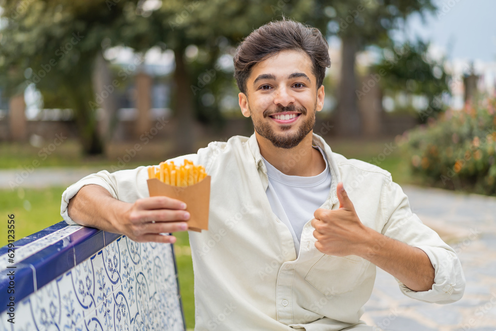 Fototapeta premium Young Arabian handsome man holding fried chips at outdoors with thumbs up because something good has happened