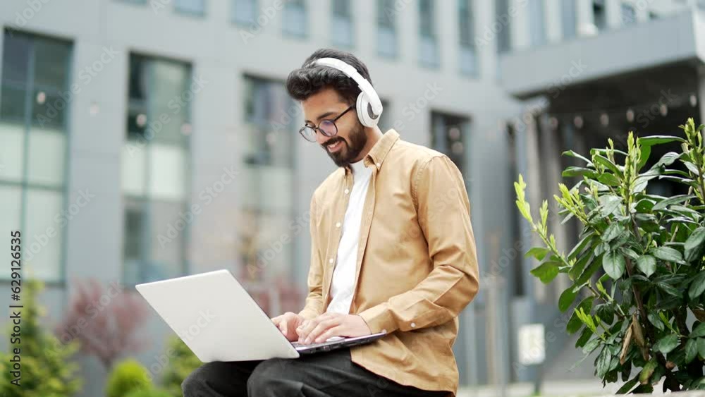 Young employee in headphones works on a laptop while sitting on a bench near the office building. Smiling handsome freelancer is chatting online with a client. IT specialist programmer uses a computer Young employee in headphones works on a laptop while sitting on a bench near the office building. Smiling handsome freelancer is chatting online with a client. IT specialist programmer uses a computer