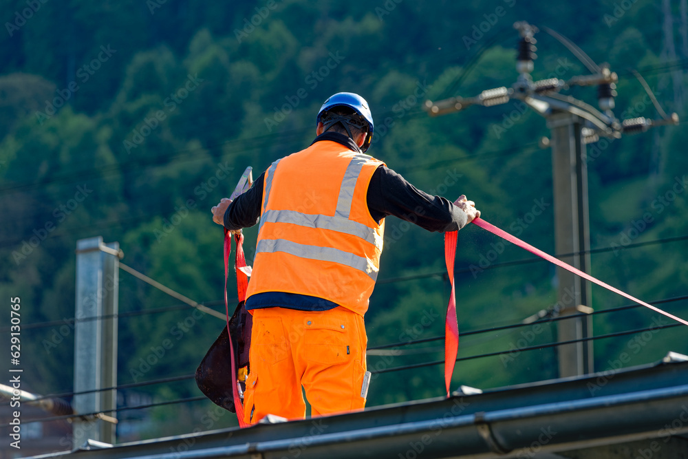 Male worker with high visibility clothing and helmet holding security belt on rooftop of platform of railway station at Swiss village of Flüelen. Photo taken May 22nd, 2023, Flüelen, Switzerland. Male worker with high visibility clothing and helmet holding security belt on rooftop of platform of railway station at Swiss village of Flüelen. Photo taken May 22nd, 2023, Flüelen, Switzerland.