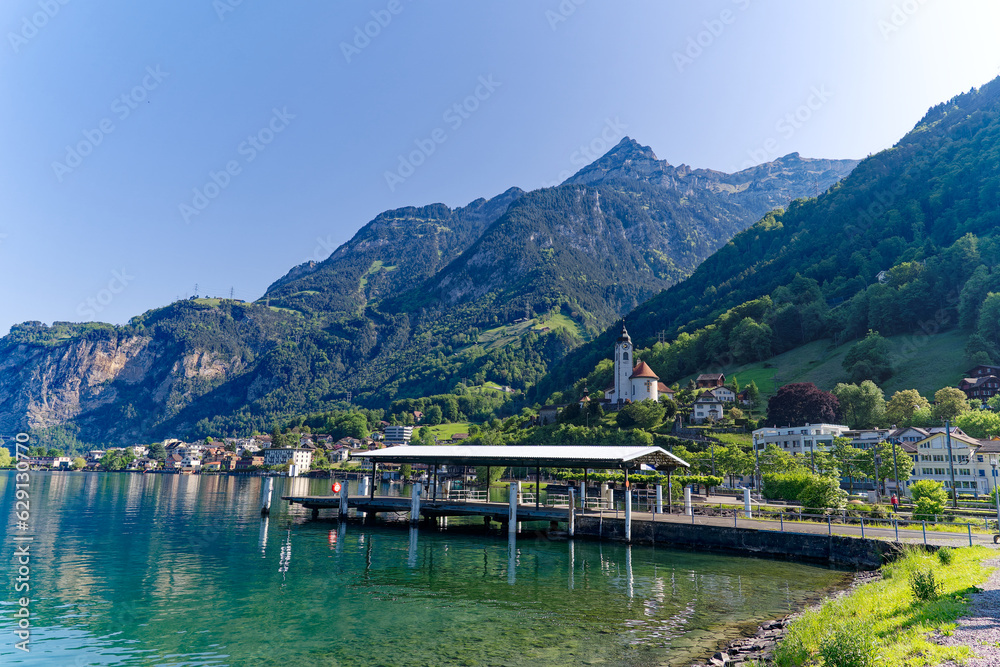 Scenic view of Swiss village of Flüelen with church tower, pier and train on a sunny spring morning. Photo taken May 22nd, 2023, Flüelen, Canton Uri, Switzerland.