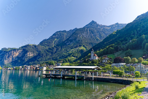 Scenic view of Swiss village of Flüelen with church tower, pier and train on a sunny spring morning. Photo taken May 22nd, 2023, Flüelen, Canton Uri, Switzerland.