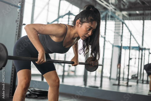 Strong Asian woman doing exercise with barbell at cross fit gym. Athlete female wearing sportswear workout on grey gym background with weight and dumbbell equipment. Healthy lifestyle.