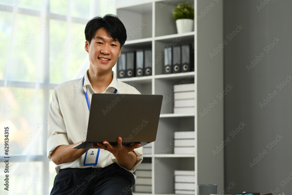Portrait of Asian male manager sitting on desk in modern workplace and using laptop computer Portrait of Asian male manager sitting on desk in modern workplace and using laptop computer