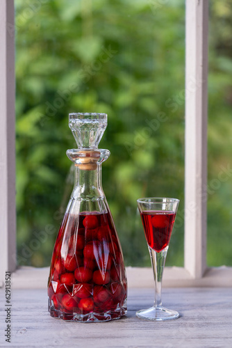 Homemade cherry brandy in wine glass and in a glass bottle on a wooden windowsill near summer garden, closeup