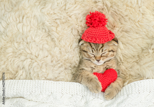 Cute fold tabby kitten wearing warm hat covered warm plaid hugs red heart on the bed at home. Valentines day concept. Top down view. Empty space for text