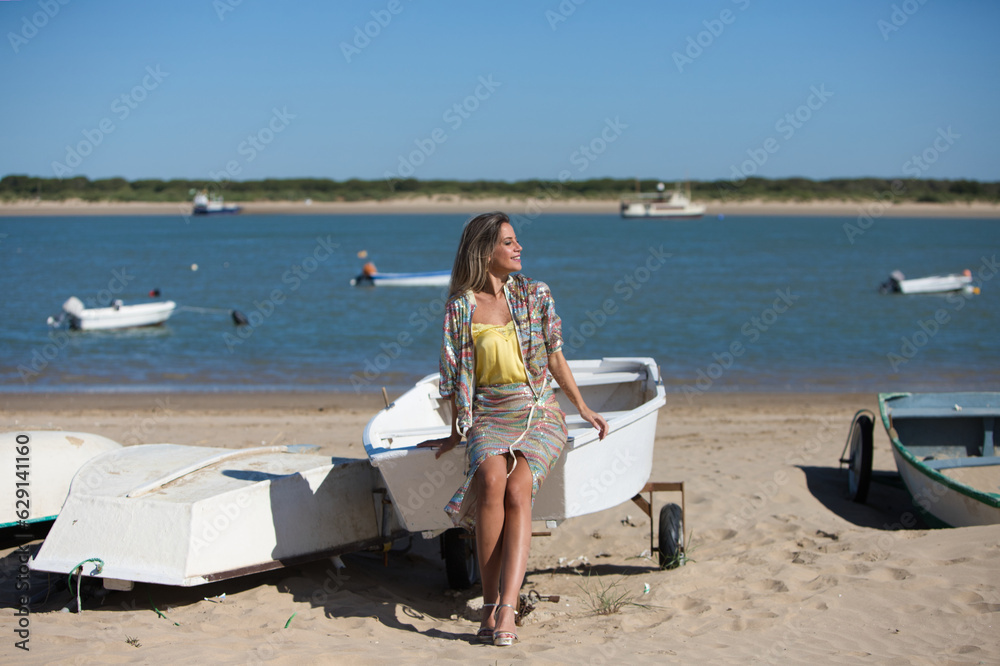 Pretty young blonde woman sitting in the fishermen's boats on the seashore. In the background on the horizon the blue sea and the boats working in the sea. Holiday and travel concept.