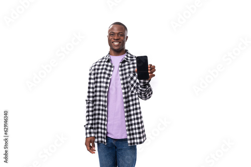 A 30-year-old African guy dressed in a plaid black and white shirt demonstrates the screen of a smartphone against the background with copy space