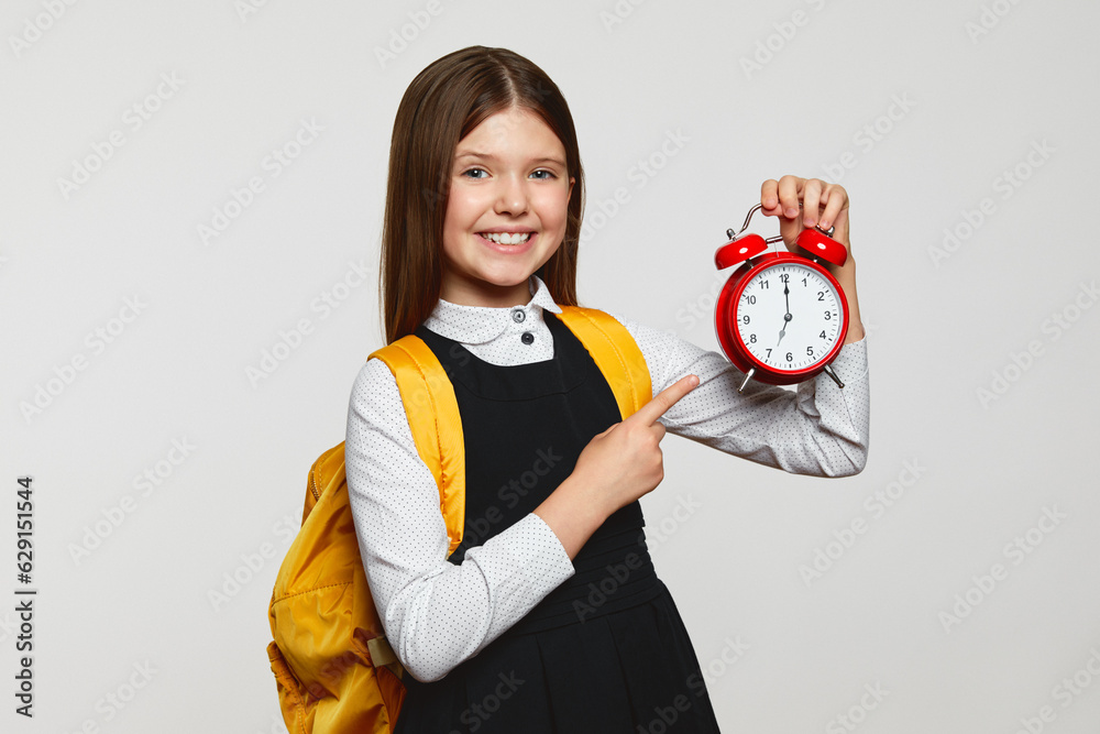 Φωτογραφία Cheerful nerdy girl in school uniform and yellow backpack holding and pointing at red alarm clock against white background
