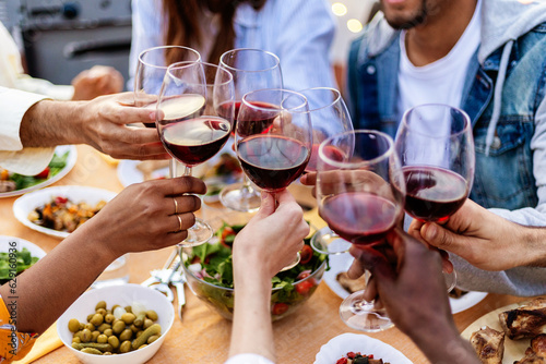 Close-up view of multiracial group of people reunited at summer party toasting red wine together. Outside evening garden celebration with happy friends enjoying dinner barbecue.