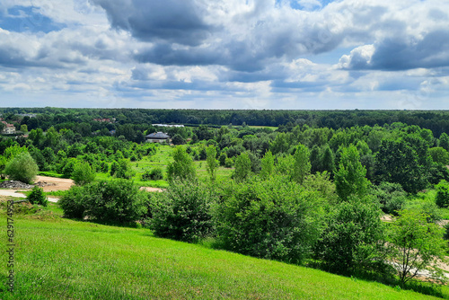 Country landscape. Rural tourism. View from the hill to houses in forest against background of cloudy sky on summer day. European countryside, Poland. Concept of environmentally friendly city.