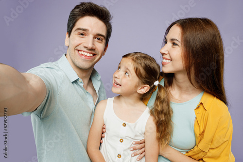 Close up fun young parents mom dad with child kid daughter girl 6 year old wear blue yellow casual clothes do selfie shot pov mobile cell phone isolated on plain purple background Family day concept.