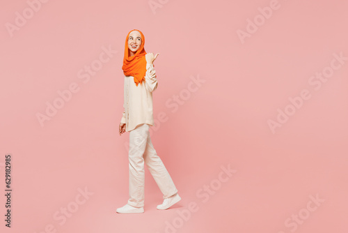 Full body side profile view young arabian asian muslim woman wear orange abaya hijab walk go strolling point finger aside isolated on plain pink background. Uae middle eastern islam religious concept.